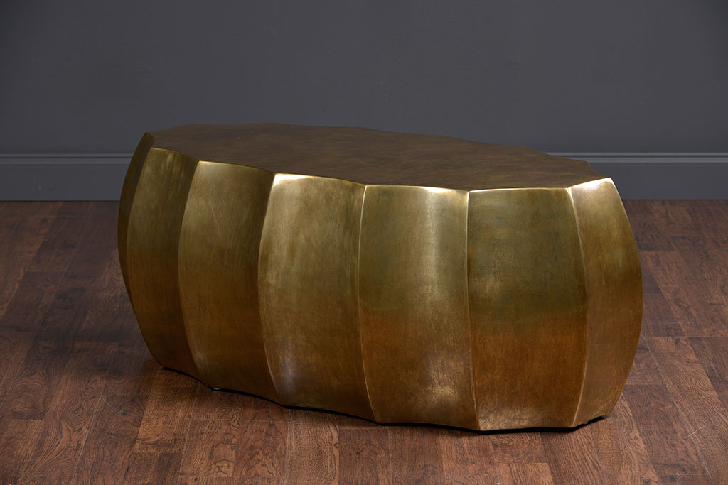 A contemporary scalloped antiqued brass oval coffee table in a room with wooden flooring and a dark wall.
