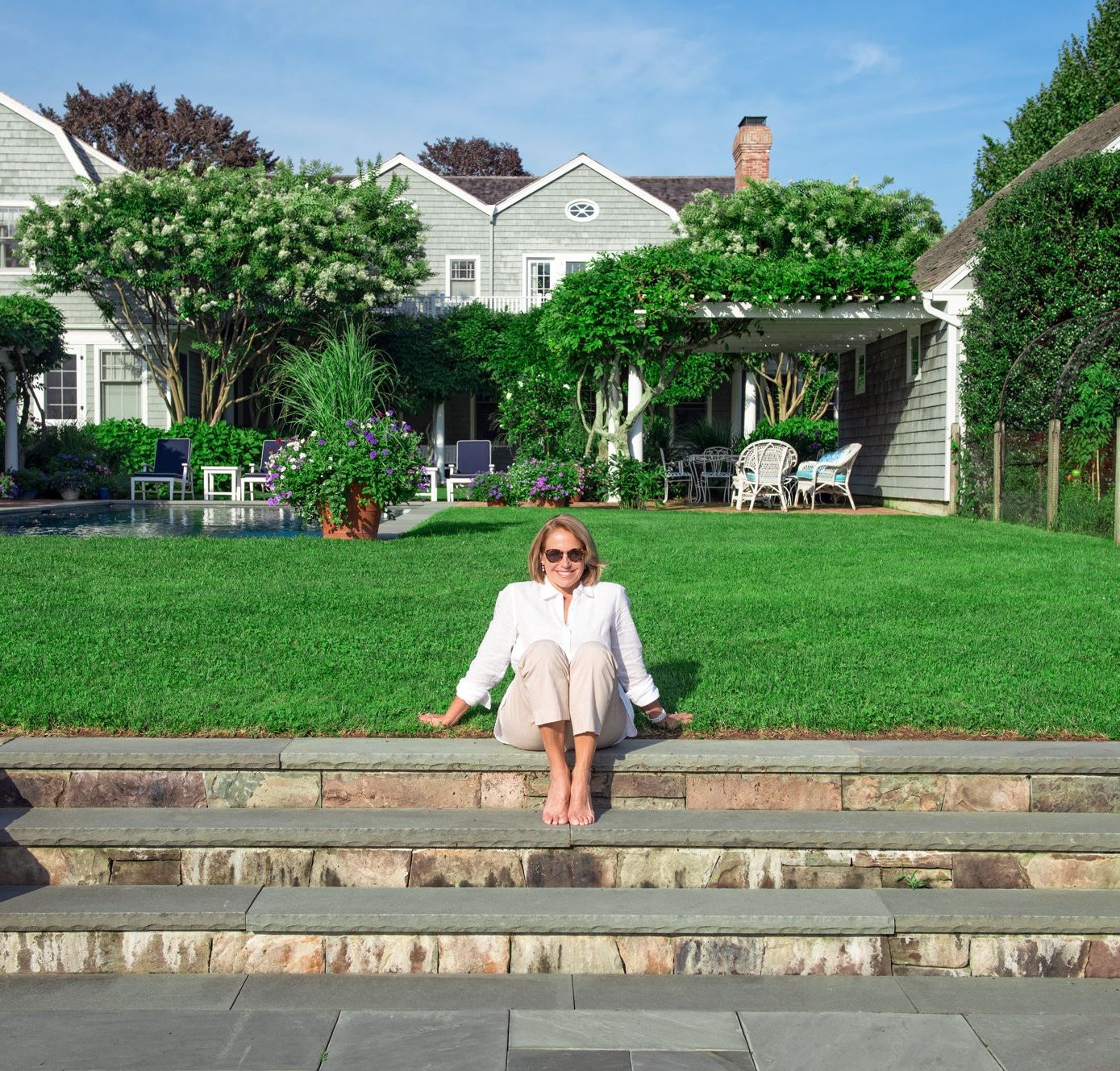 Woman sitting on stone steps in front of a large house with white siding and multiple windows wearing sunglasses and a light-colored