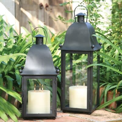 Two black lanterns with candles, placed on a patio surrounded by green foliage.
