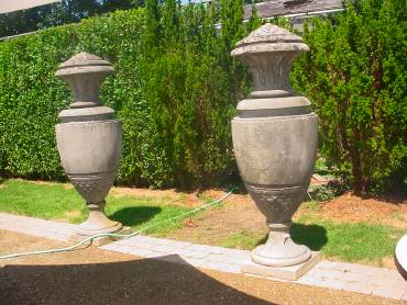 Two ornate stone vases in a garden, set against a backdrop of lush greenery and a clear sky.