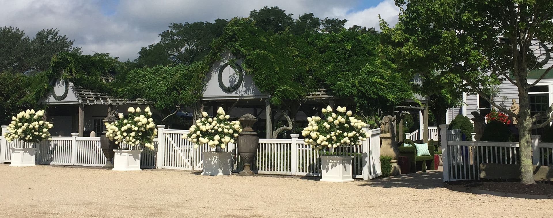 White gazebo with a black roof, surrounded by potted plants and flowers, set against a backdrop of trees and sky.