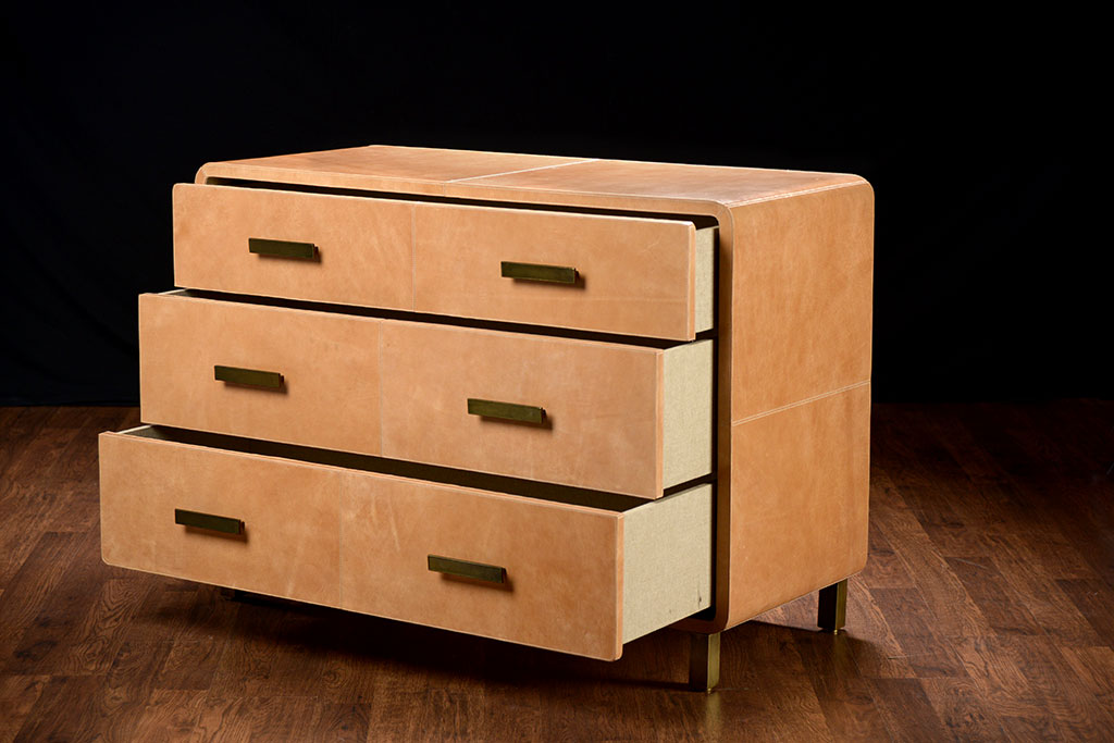 Brown leather dresser with curved edges and three drawers, one of which is open, set against a dark background.