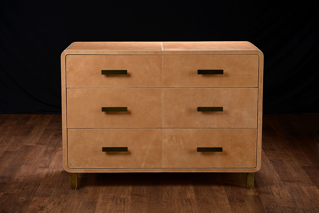 Brown leather dresser with three drawers, curved edges, and gold metal feet on a dark background.