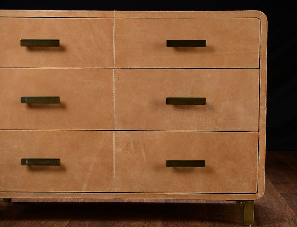 A brown leather dresser with three drawers a curved front design and gold metal feet The dresser is part of a set
