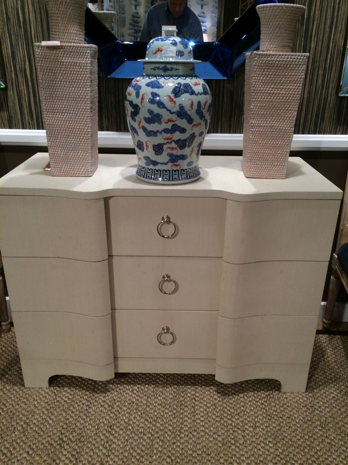 A large white dresser with a blue and white vase on top, featuring silver ring pulls and a grasscloth finish.