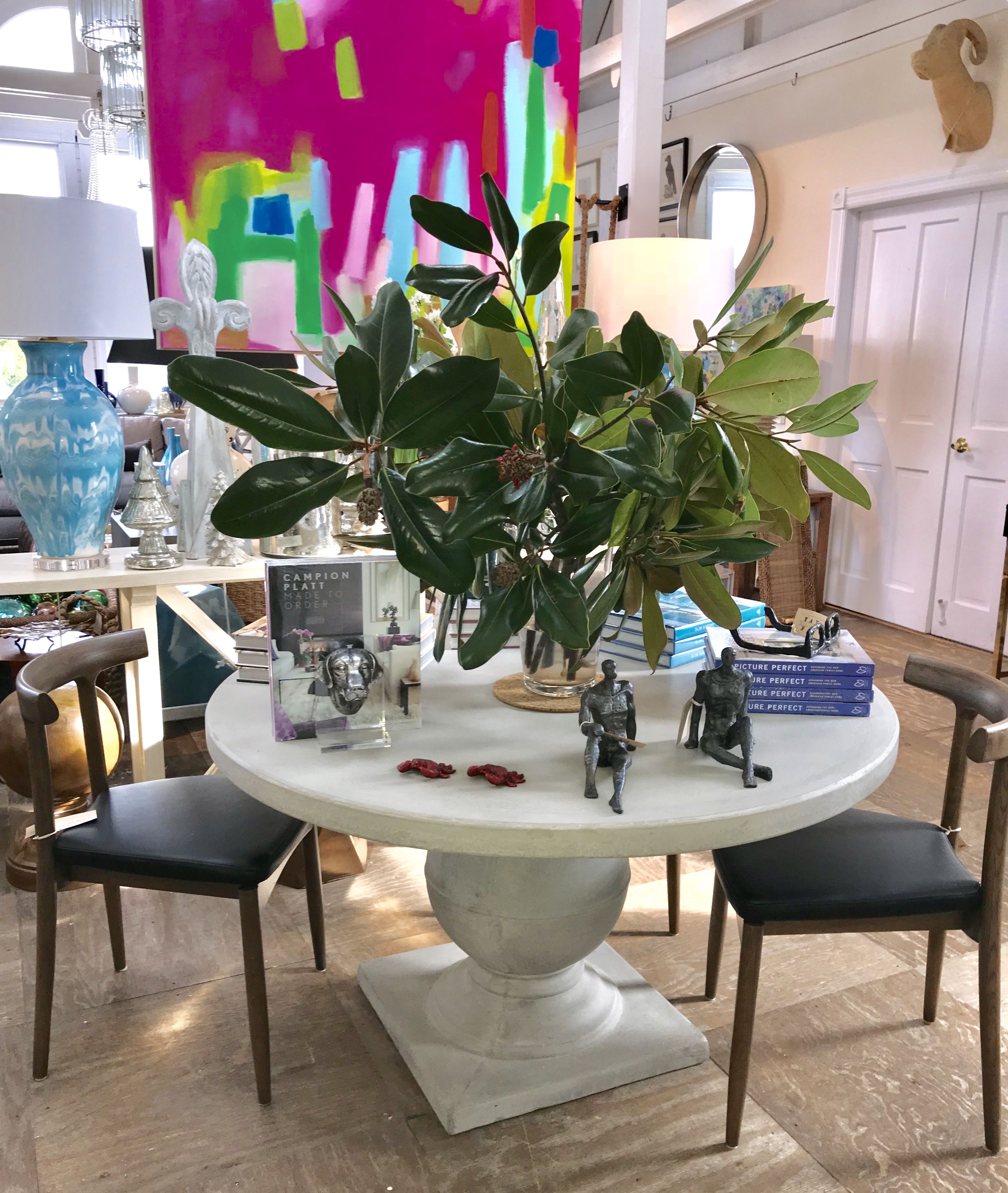 A round concrete dining table with a light gray finish set against a backdrop featuring a colorful painting and a potted plant
