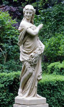 Statue of a maiden in classical attire, carved from stone, with flowers at her feet, set against a garden backdrop.